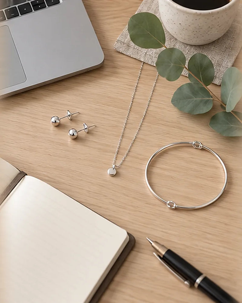 Dezenter Büro-Schmuck: Silberohrstecker, feine Kette und schlichtes Armband neben Laptop und Notizbuch